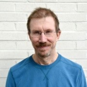 Bildet er et portrettfoto av Petter Holm med blå skjorte foran en hvit vegg