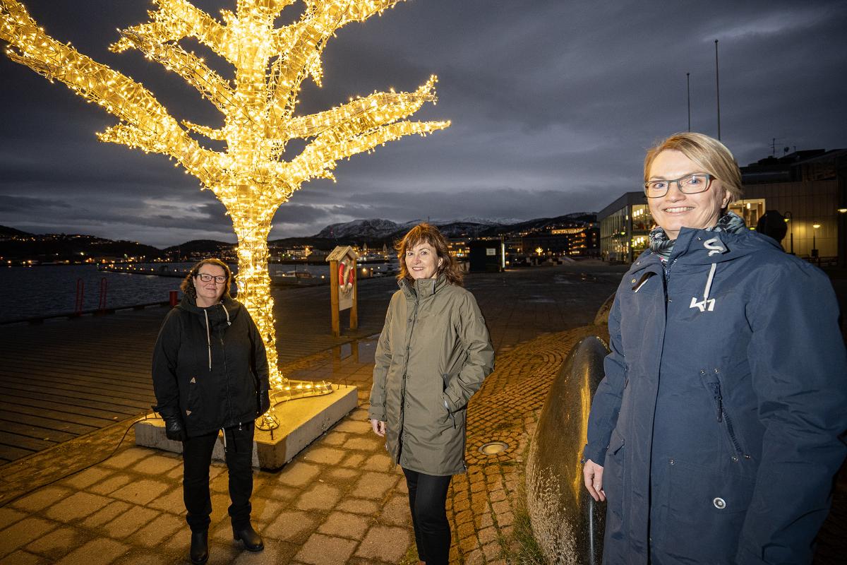 3 damer står utendørs foran et julebelyst tre