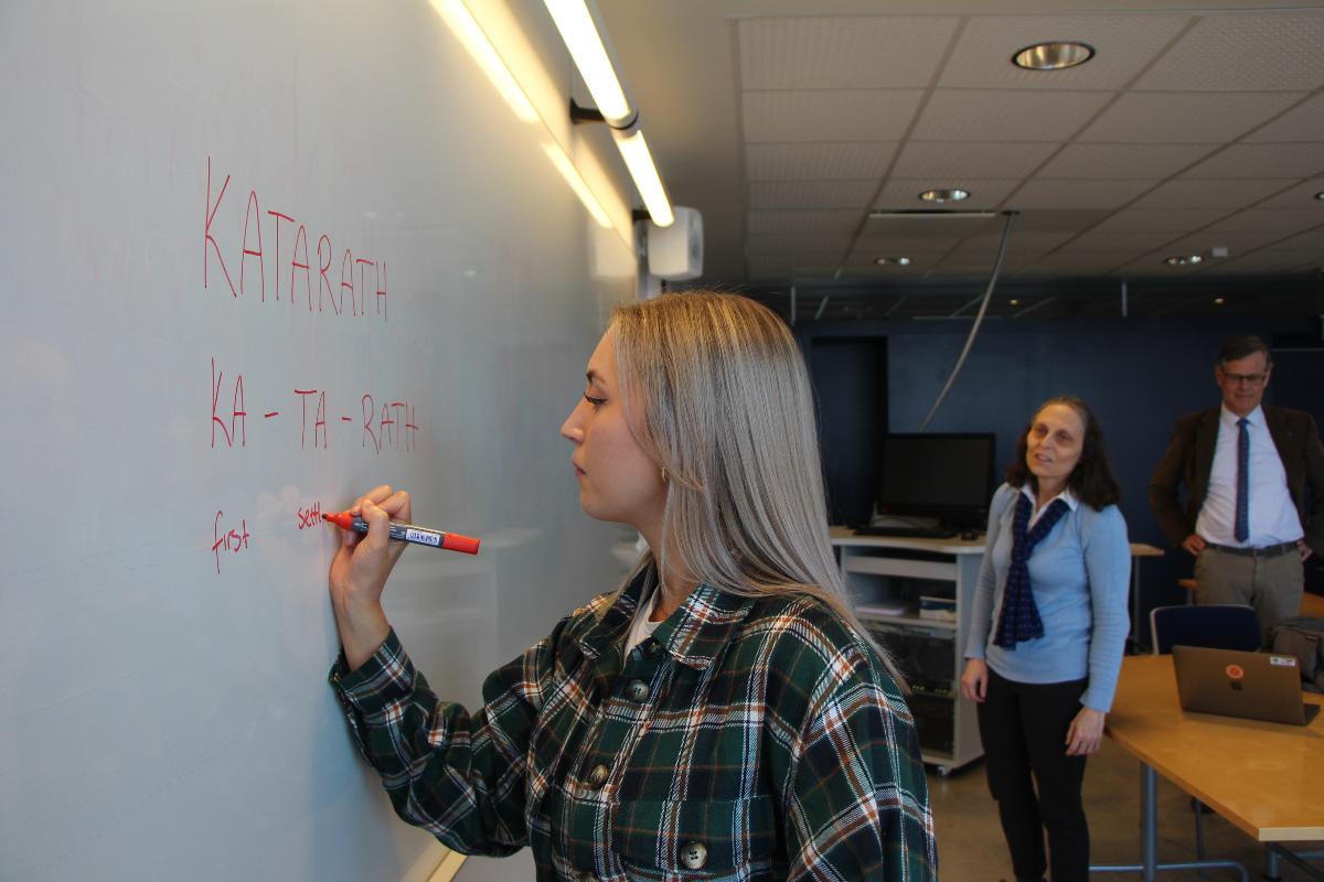 Ung kvinne, Marte Isaksen, skriver på tavla med entusiastiske lærere i bakgrunnen (professor Laura Janda og Tore Nesset)
