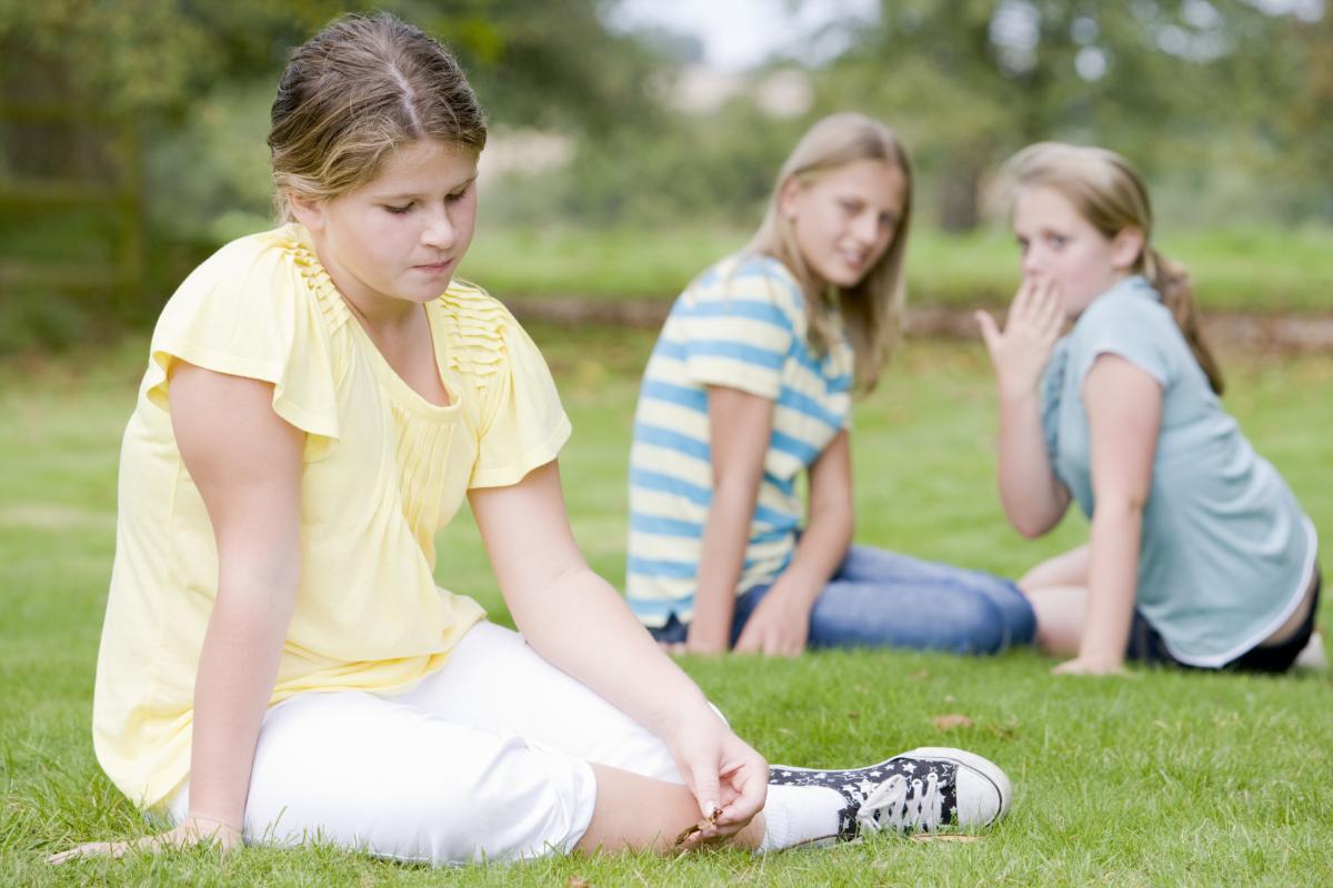 To jenter utestenger en tredje jente, ute på en gressplen. De hvisker og ser på den tredje jenta.