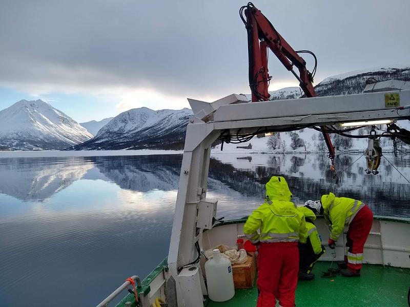 Tre personer på dekk av en båt, fjord og snødekte fjell i bakgrunnen.