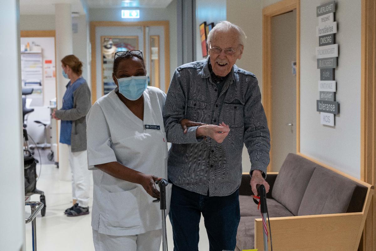 Smilende gammel mann og sykepleier i gangen på sykehjem