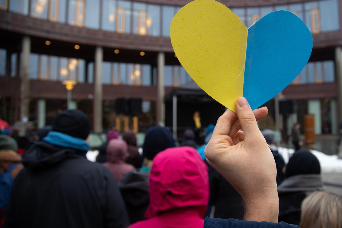 Hender som holder et hjerte med farger fra det ukrainske flagget foran et folkehav. 