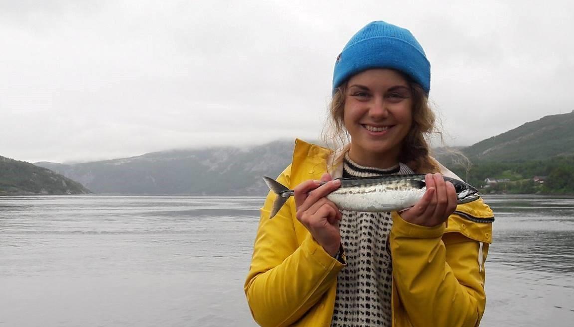 Bilde av Mathilde, mens hun holder en fisk. En fjord er i bakgrunnen av bildet. 