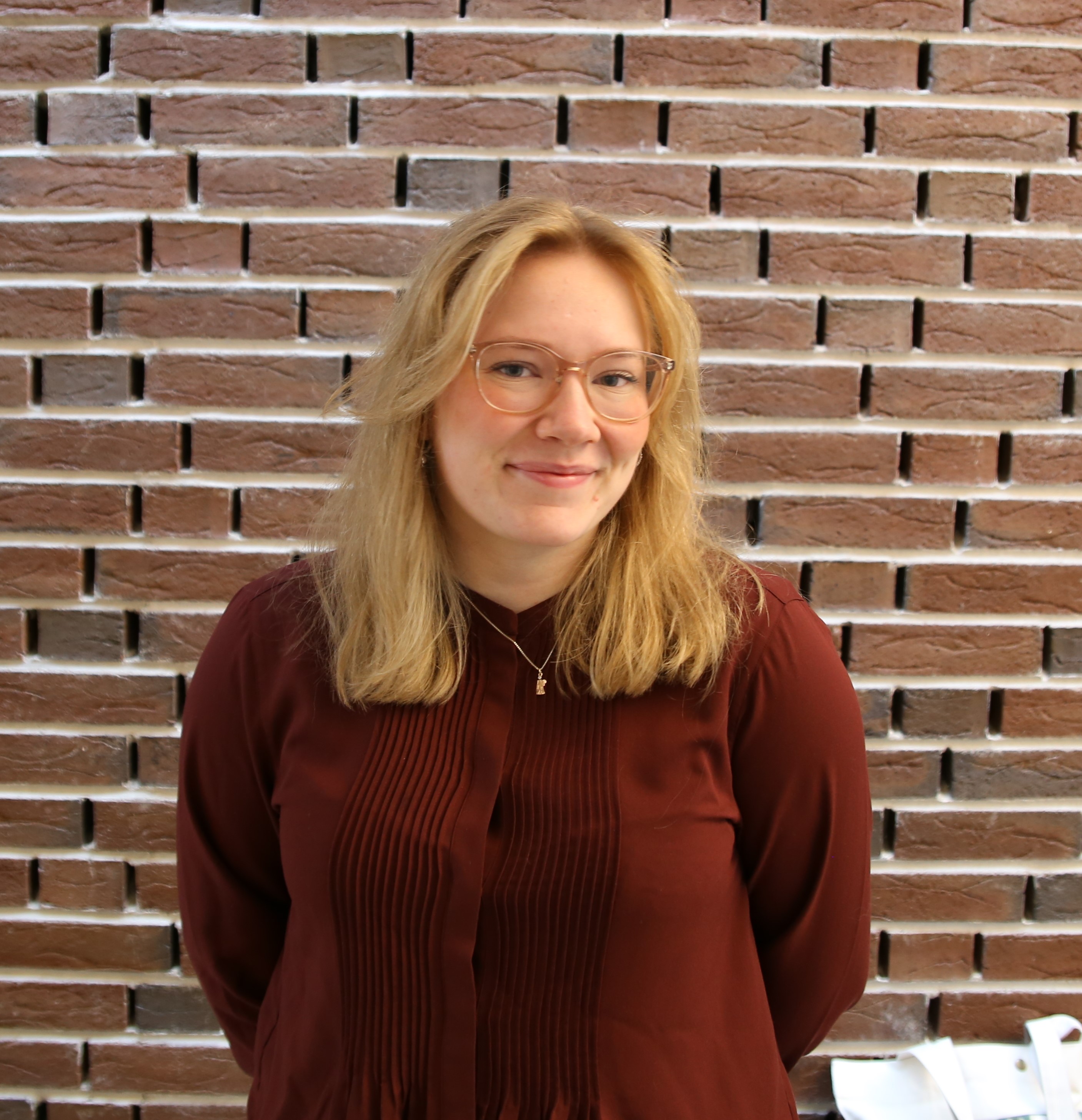 Bilde av Ida Jørgine i profil. Hun har blondt hår, briller og en mørkerød skjorte på seg. I bakgrunnen er en murvegg.