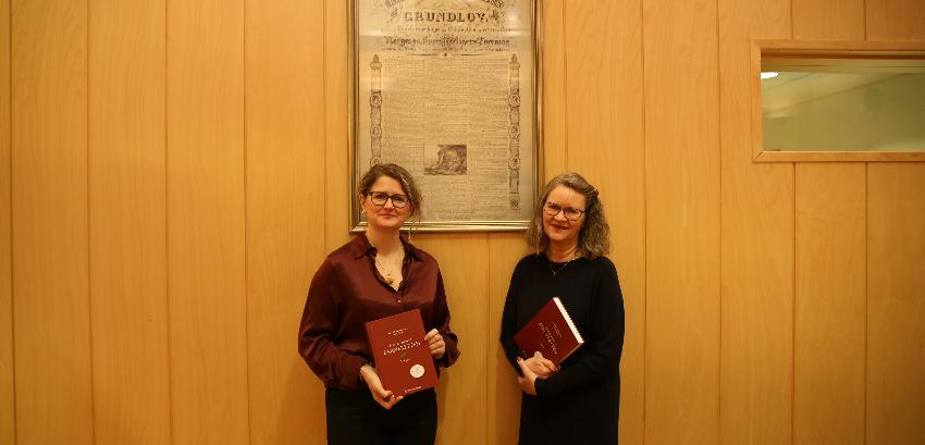 Forfatterne poserer med boka foran en poster av Grunnloven. 