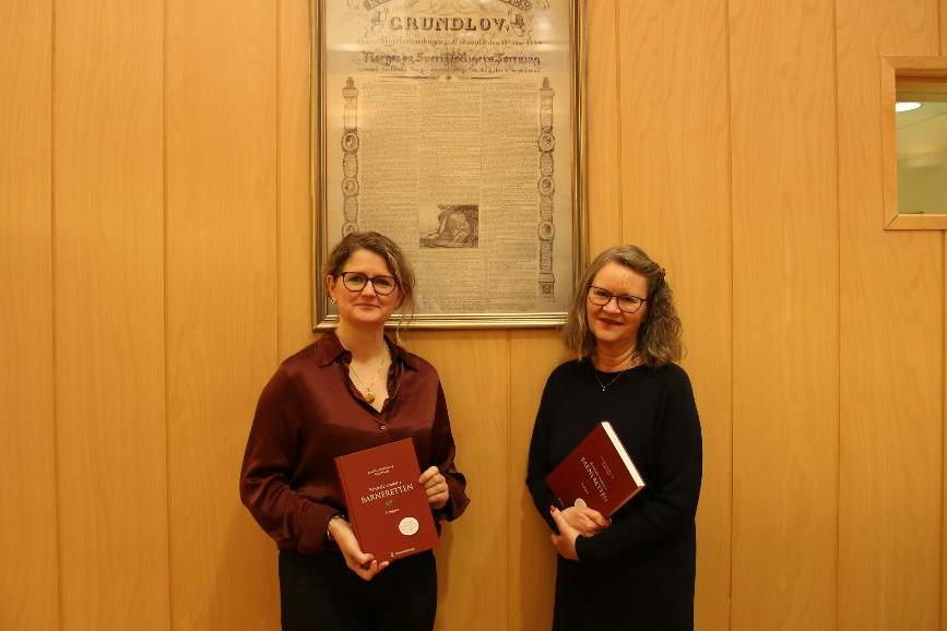 Forfatterne poserer med boka foran en poster av Grunnloven. 
