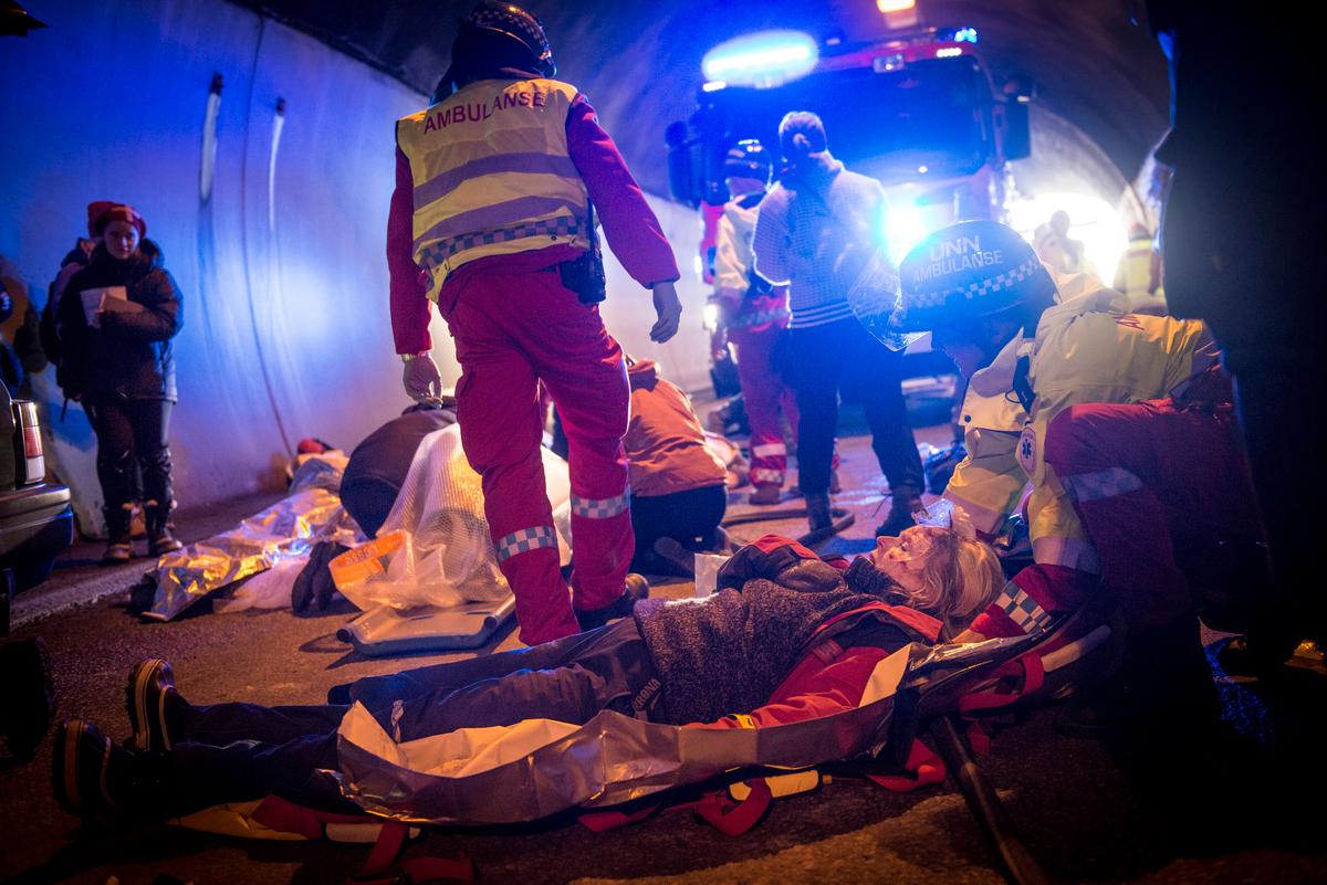 Kaotisk scene frå ein tunnel. Skadde ligg på bakken, ein ambulansearbeidar står med ryggen til. Brannbil med blålys i bakgrunnen.