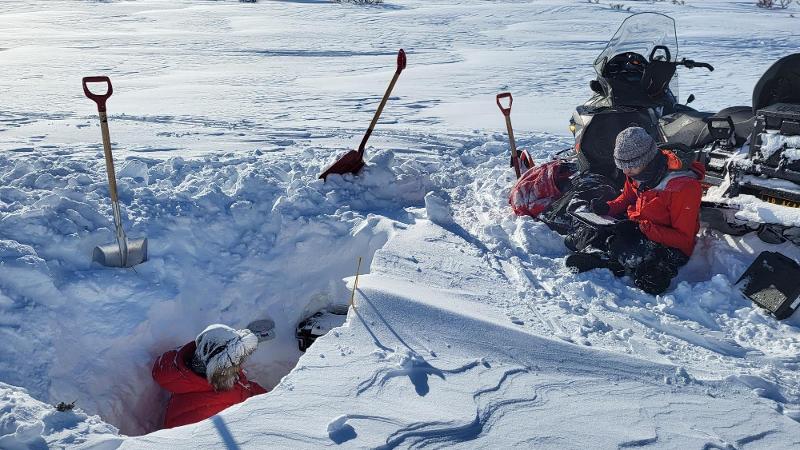 EN person er nede i et hull i et snølandskap. ovenfor hullet er en annen person, en snøskuter og mengder med utstyr