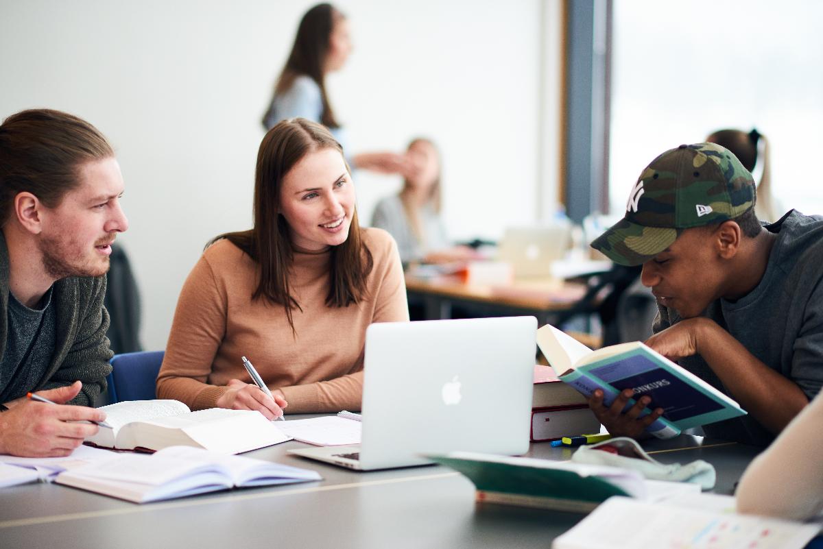 Tre unge mennesker rundt et bord, med b&oslash;ker og PC