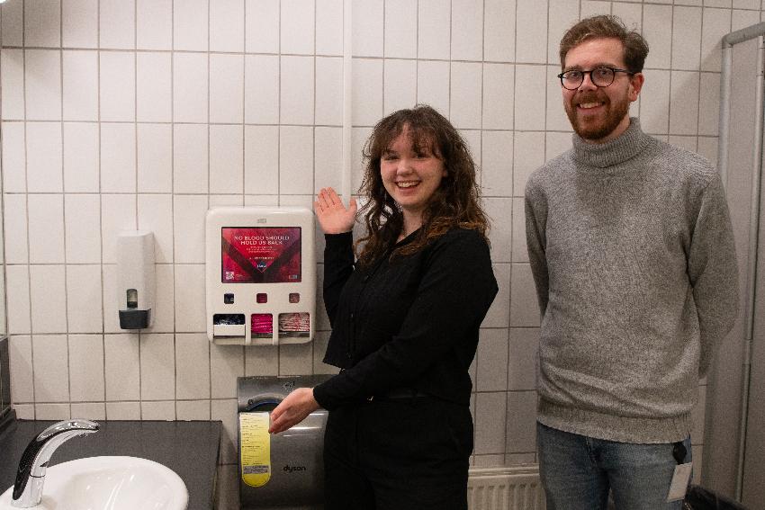 En ung kvinne og ung mann poserer med en sanitærdispenser på et toalett