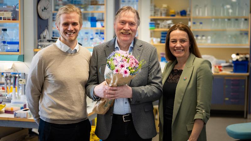 To menn og en kvinne poserer i et laboratorium. Mannen i midten holder en blomsterbukett.