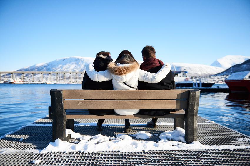 tre personer sitter med ryggen til på en benk ved tromsø havn. En bro, et sund og snødekte fjell i bakgrunnen.