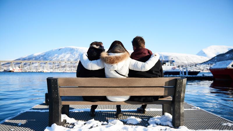 tre personer sitter med ryggen til på en benk ved tromsø havn. En bro, et sund og snødekte fjell i bakgrunnen.