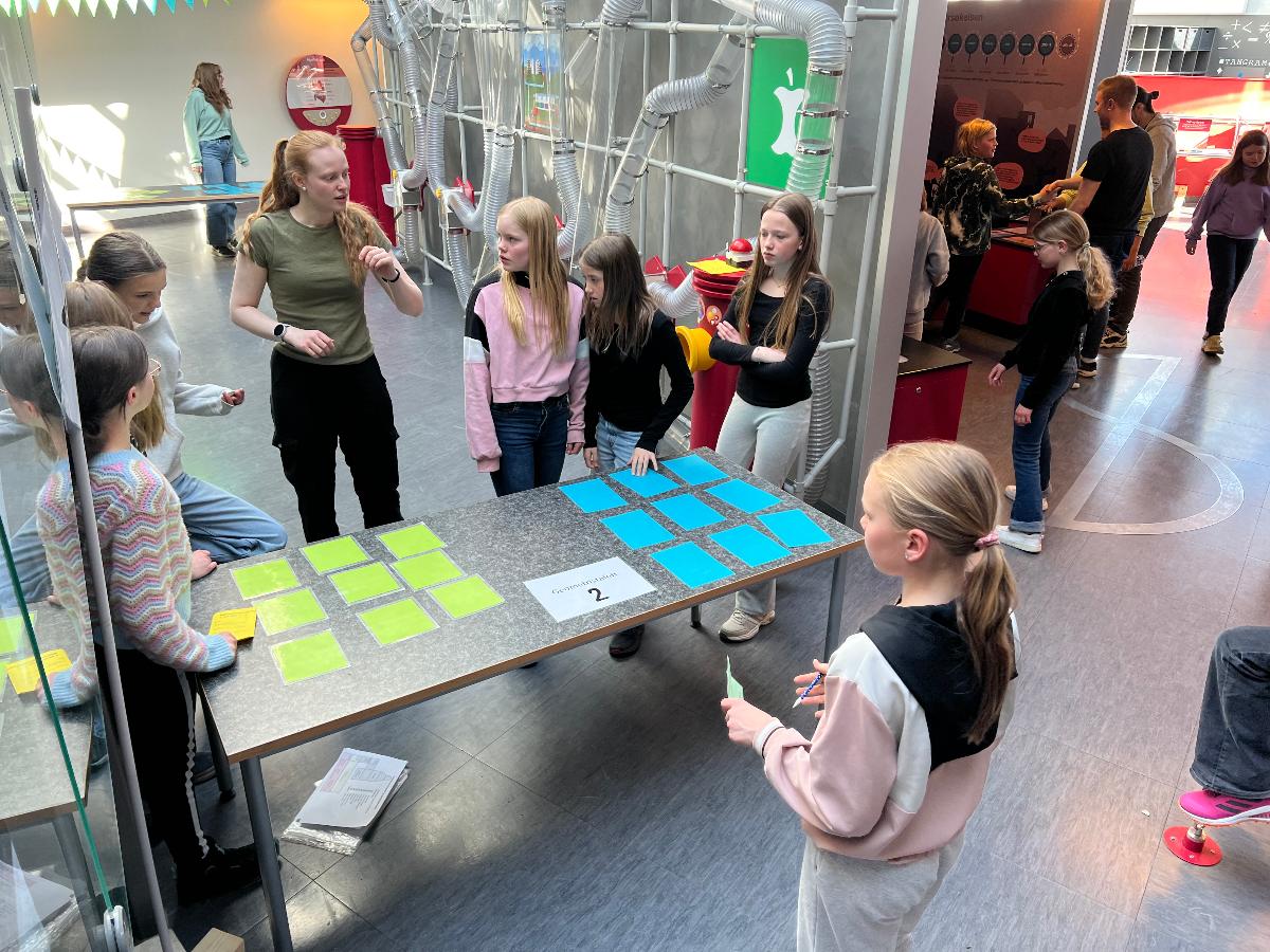 Åtte elever i sjetteklasse får hjelp av studentassistent til å løse en praktisk matematikkoppgave. Stort lokale med bord med lapper i blå og gul farge.