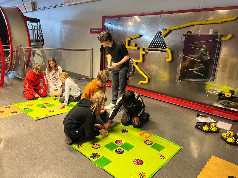 Seks sjetteklassinger sitter på gulvet foran en labyrint med en bie-robot som de skal programmere til å fly riktig vei i labyrinten.