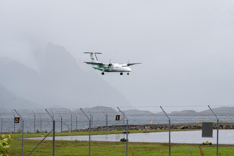 Et hvitt og grønt fly inn for landing i ruskevær. Opprørt sjø og fjell i bakgrunnen.