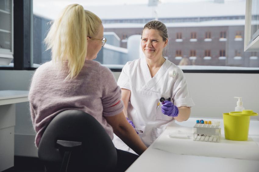 Kvinne i sykehusklær smiler til en person med ryggen til. De sitter i et laboratorium ved et stort vindu.