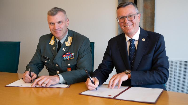 En mann i grønn uniform og en mann i dress sitter ved et bord med papirer foran seg. De smiler mot kamera og holder hver sin penn.