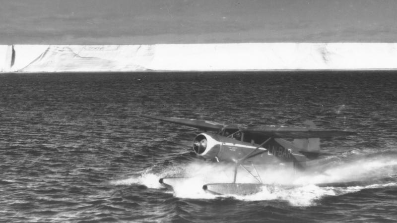 black and white photo of a sea plane landing in front of a giant glacier / ice sheet