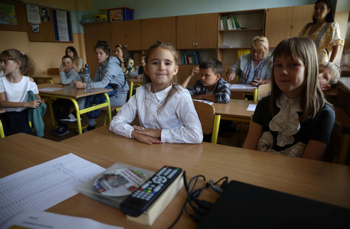 Jente med hvit bluse ser stolt inn i kamera. Hun sitter i et klasserom med andre barn og voksne.