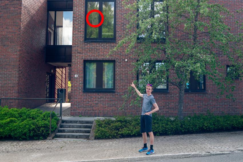 ein mann i sommerklede peiker på ein bygninga bak seg. det er teikna ein raud ring rundt ein sak festa på eit vindauge