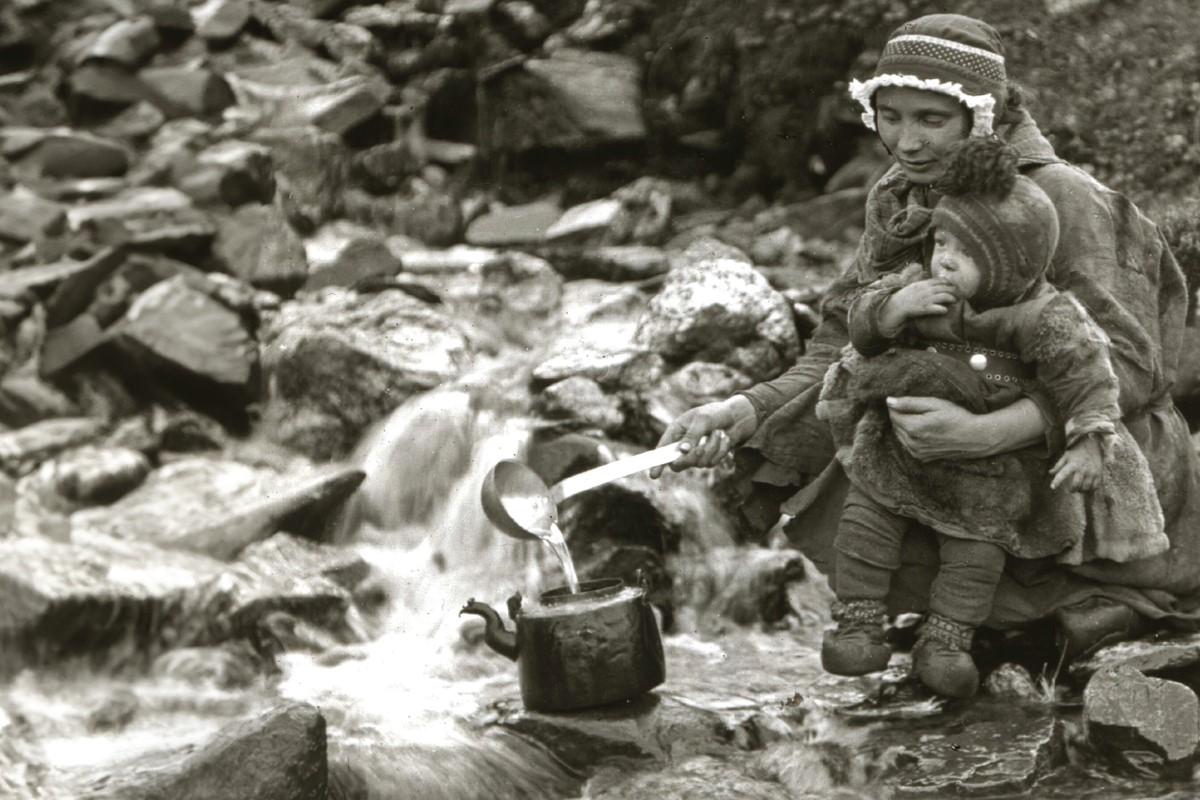  Eldre sort-hvitt foto av kvinne med spebarn som fyller vann i en kaffekjele fra en bekk