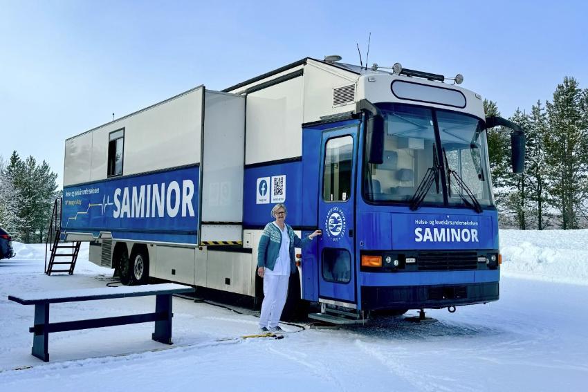 En dame i sykehusklær og ulljakke står ved en buss i vinterlandskap. På bussen står det SAMINOR.