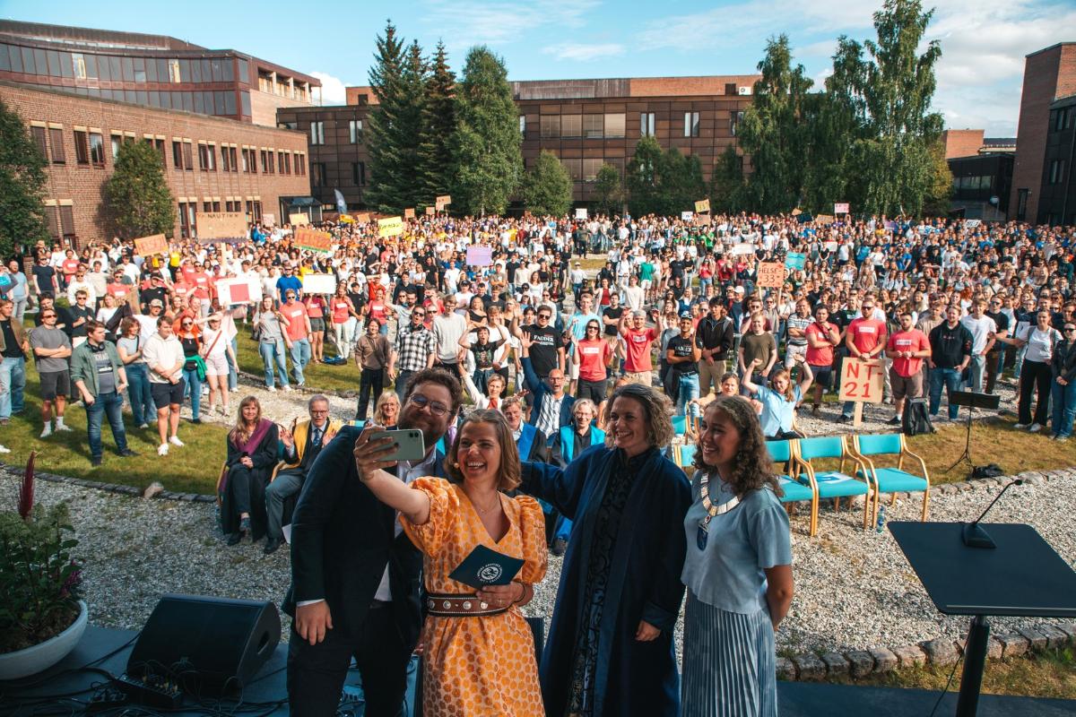 Fire ledere på en scene tar selfie med masse studenter i bakgrunnen på et et grønt og frodig universitetsområde. 