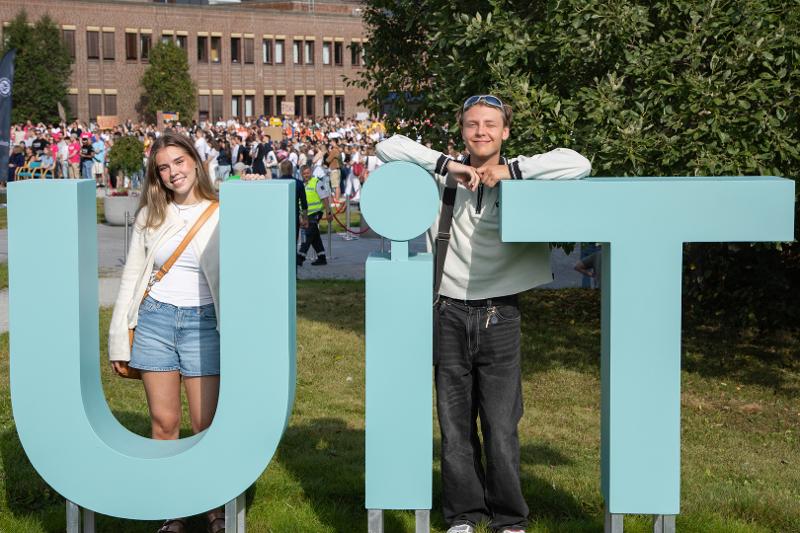 To studenter, kvinne og mann, poserer ved UiT-logo på campus.