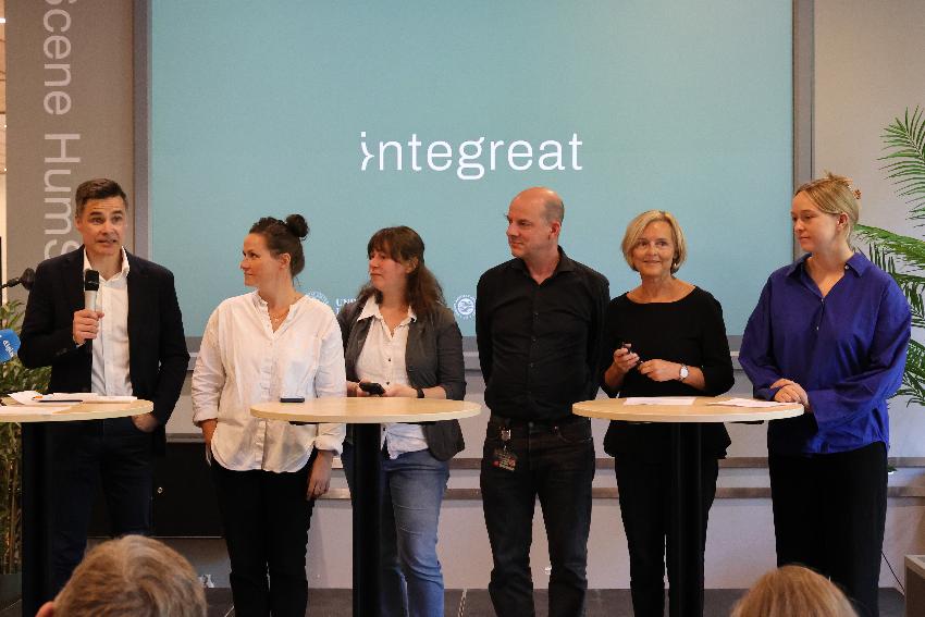 En gruppe mennesker i et panel med runde bord foran seg. Bak dem en skjerm det står "integreat" på.