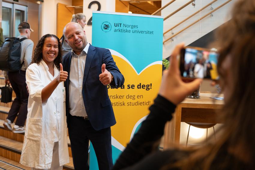 Minister og student holder sine tomler opp til videofotograf inne på et studiested med bannere og flere studenter. 