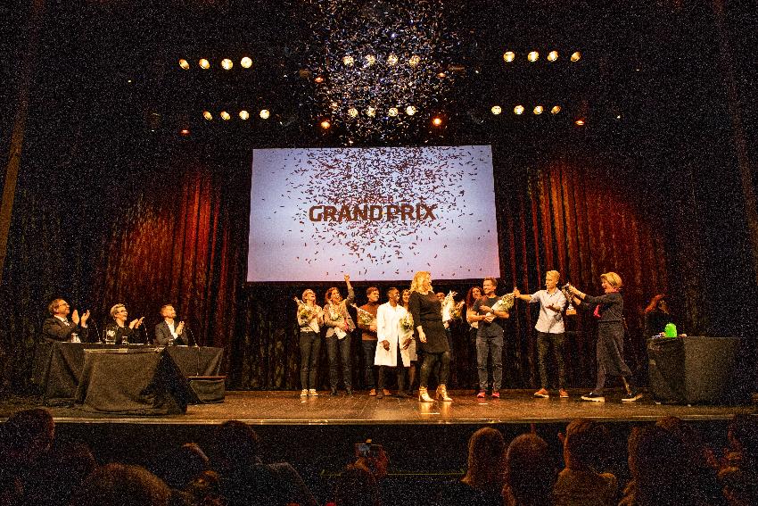 Folk på en scene smiler og klapper. En dame står foran og mottar en pokal.