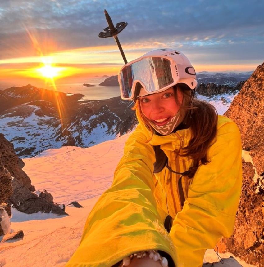 En kvinne med skiutstyr på en fjelltopp med solnedgangen i bakgrunnen.