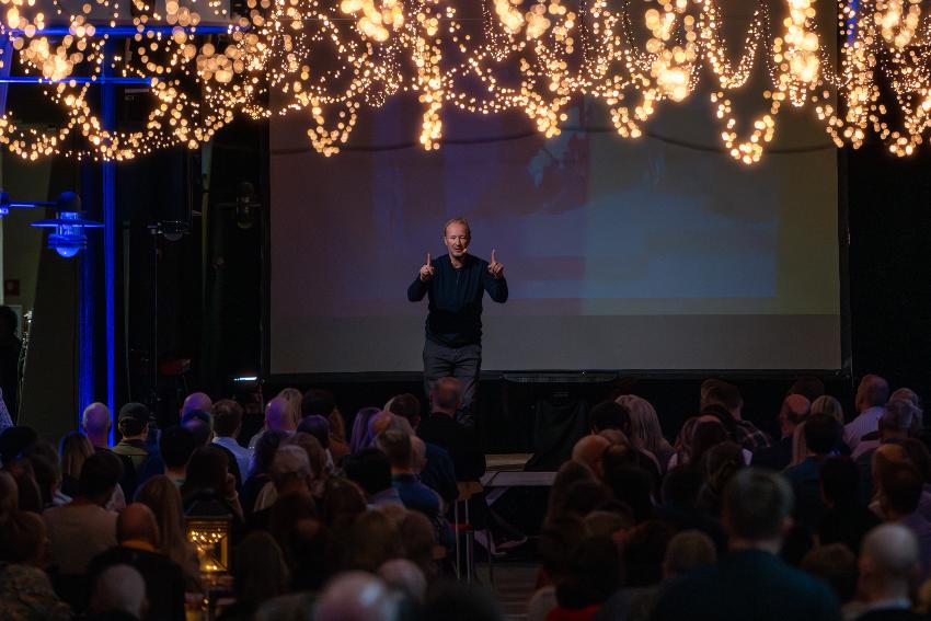 Blond mann står på en scene, gestikulerer med begge hendene i været.