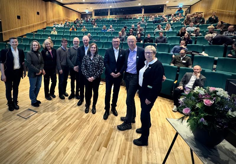 Flere personer står ved siden av hverandre foran en sal. 