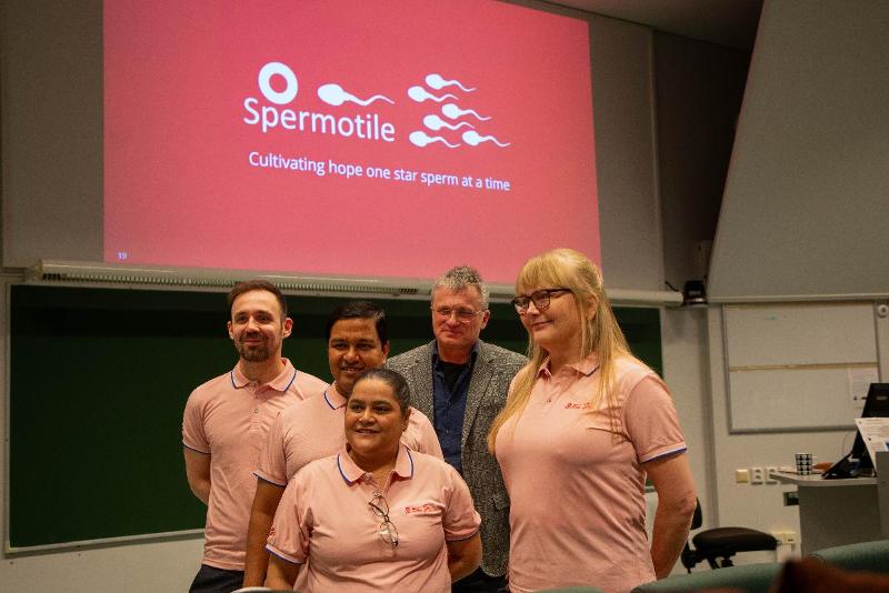 Fem personar poserar fremst i eit auditorium. Det står spermotile på skjermen bak dei.