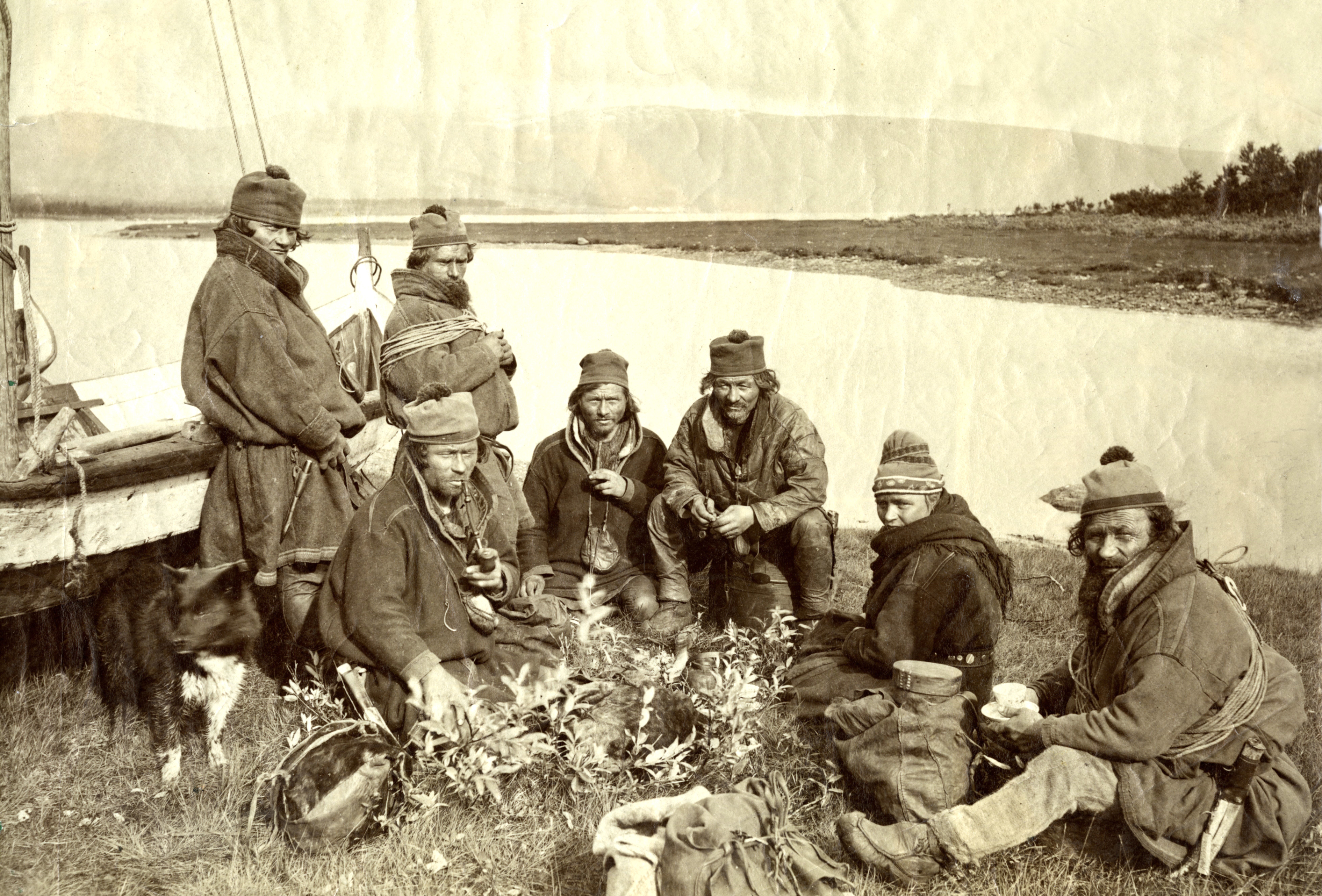 I fjæra samer som sitter i fjæra ved siden av en båt. Noen røyker pipe.