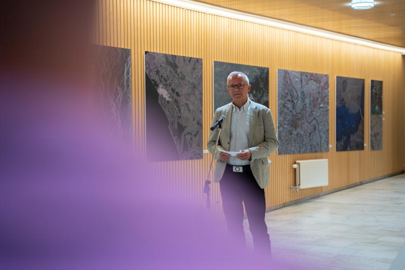 Bildet viser Kai Mortensen, medlem av kunstuvalget som holder tale. I bakgrunnen ser man bilder fra ustillingen. Mortensen har på seg en lys dressjakke og mørk bukse. 