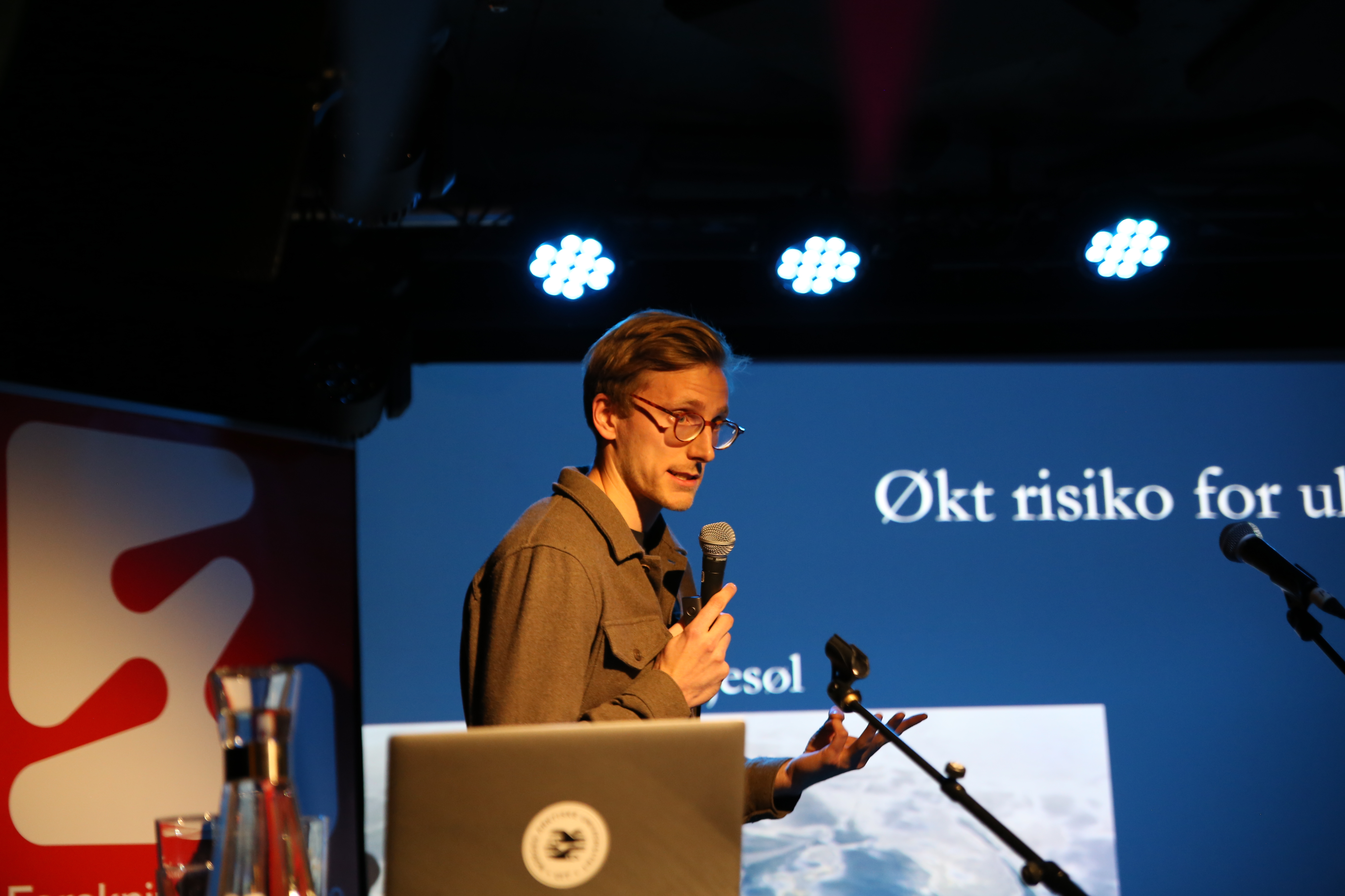 Mann ser mot publikum og snakker i mikrofon. 