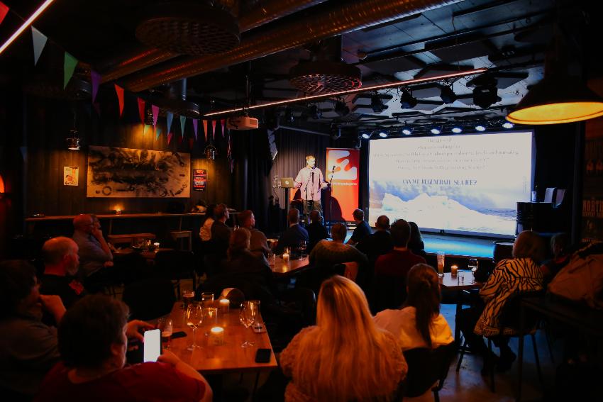 Bilde av lokalet på Amtmandens mot scenen, man ser publikum i rommet, og på scenen presenterer Apostolos Tsiouvalas. 