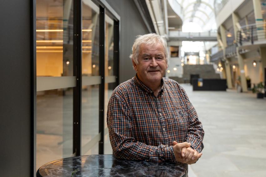 Hvithåret mann i rutete skjorte smiler til kamera, lent inntil et bord i en glassgang