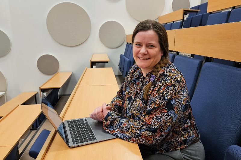 Kvinne sitter i et auditorium med pc og ser i kamera