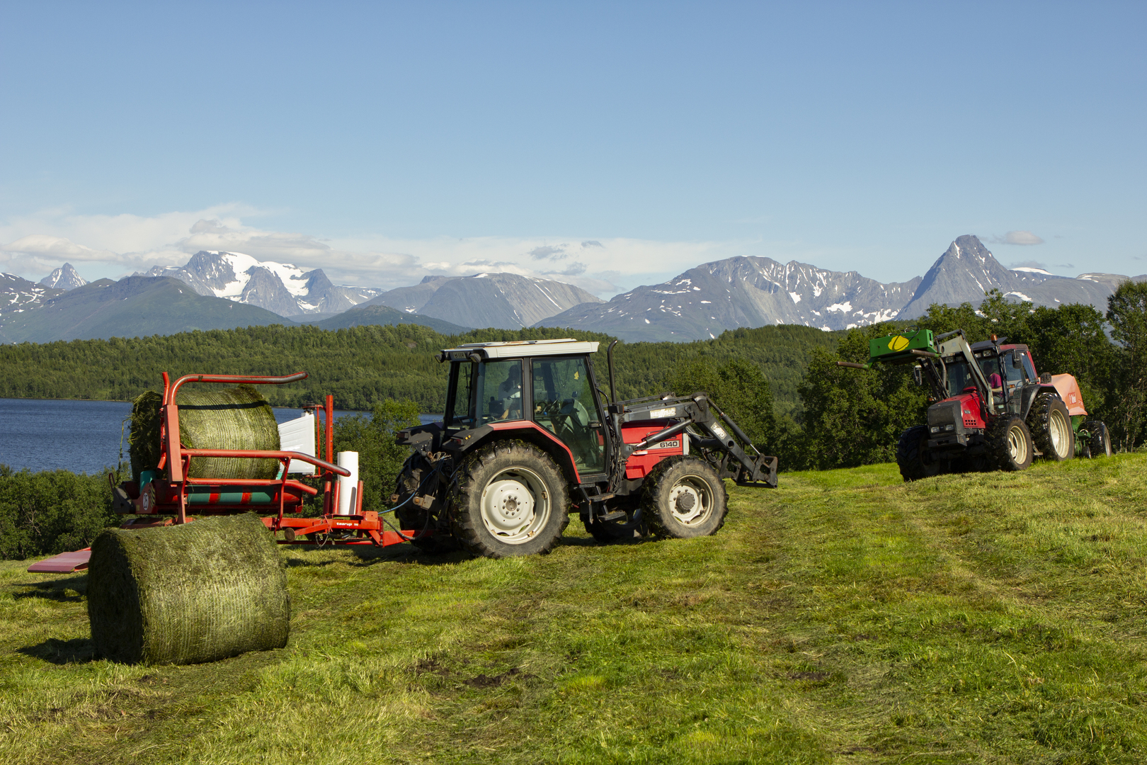 To traktorer på et jorde, med fjell og fjord i bakgrunnen.