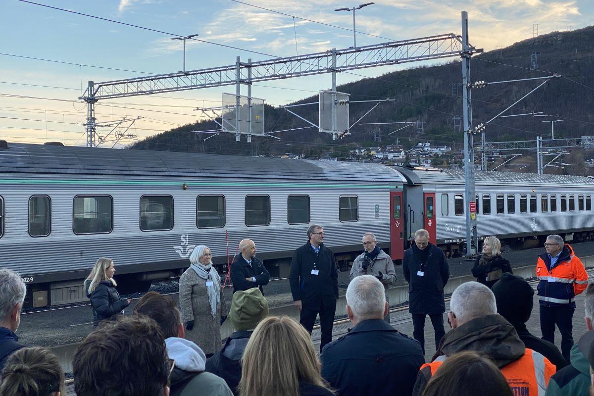 et tog står parkert og folk er samlet på perrongen mens noen holder tale