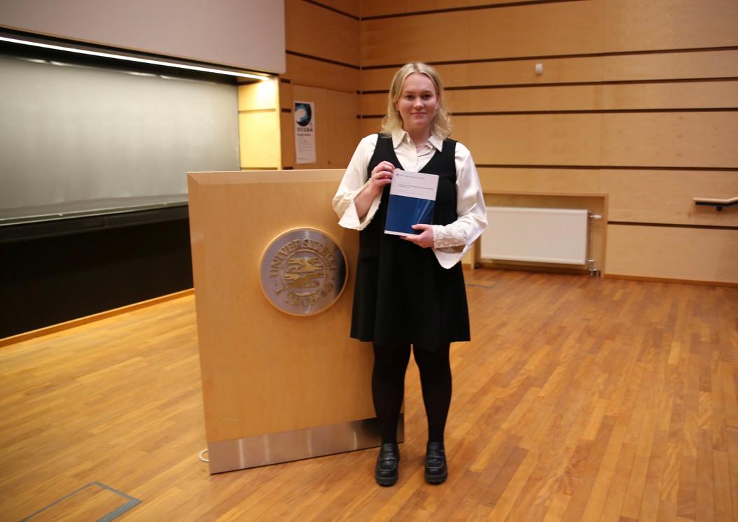 Jente blondt h&aring;r holder opp avhandling og ser inn i kamera, podium bak