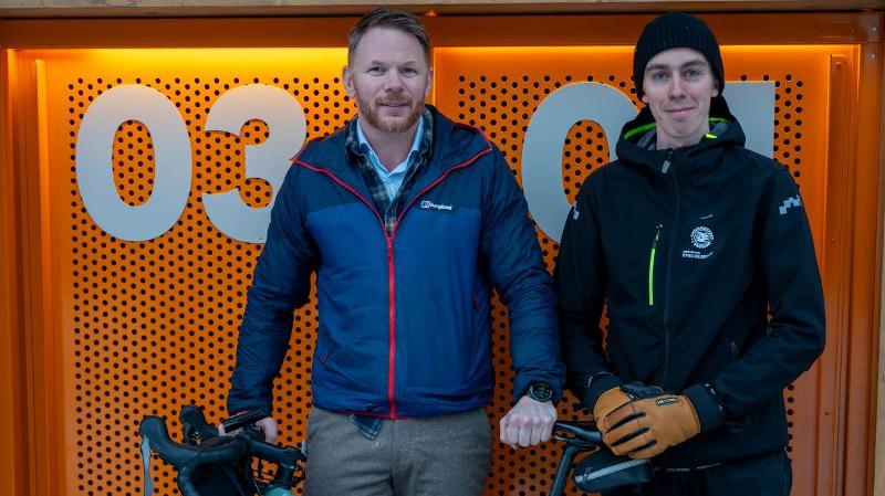 to menn som st&aring;r foran et sykkelskur med en turkis sykkel