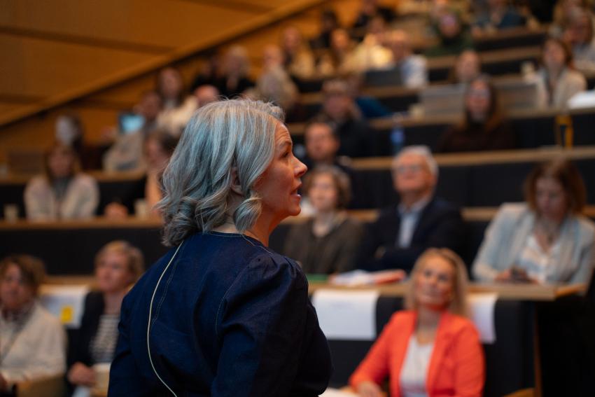 Kvinne taler til forsamling i forgrunnen. 