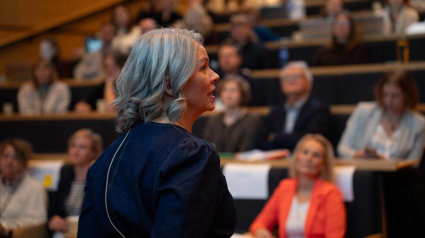 Kvinne taler til forsamling i forgrunnen.