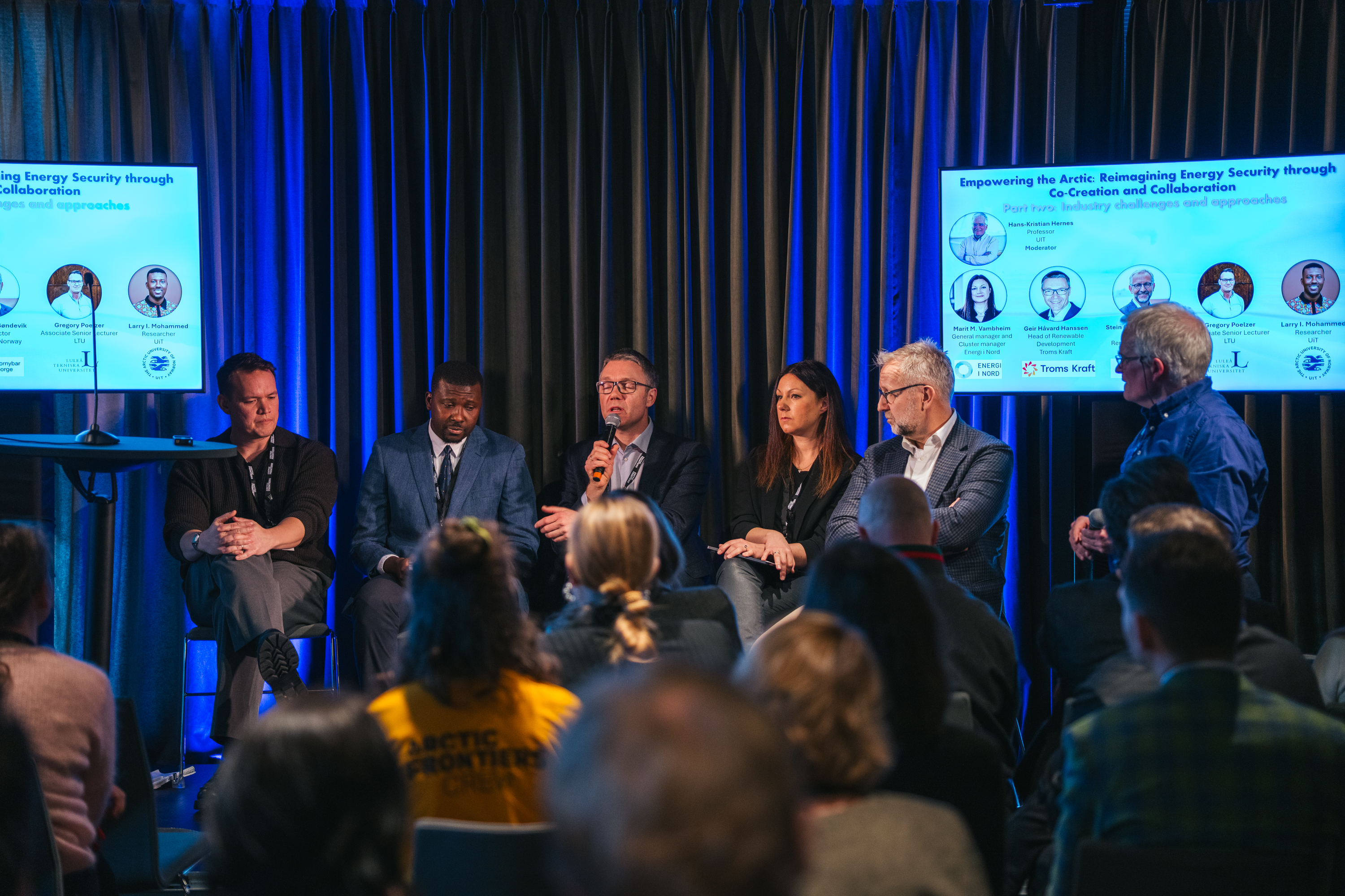 Personer som sitter p&aring; en scene mellom to skjermer.